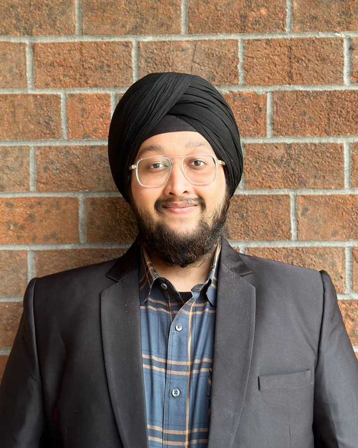 Portrait of Davindernain Singh Sehgal in a dark blazer and plaid shirt, smiling, in front of a brick wall