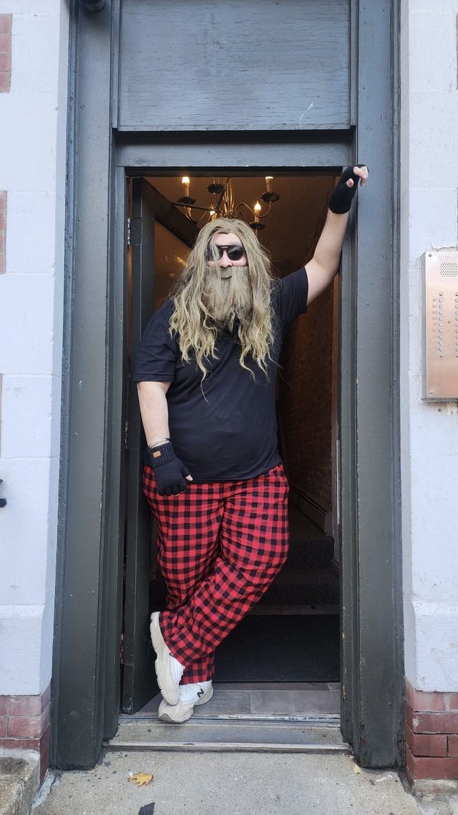 A person in a long grey wig and plaid pants standing in a doorway