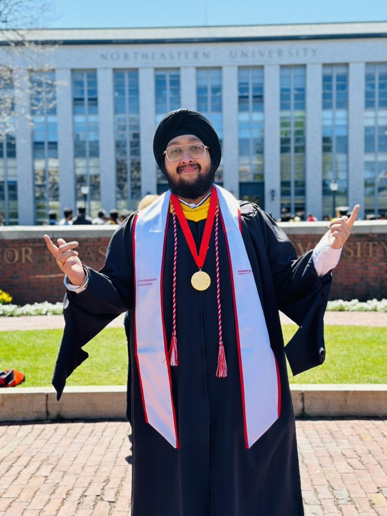 Dave in his graduation gown and honor stole, arms out, in front of a university building