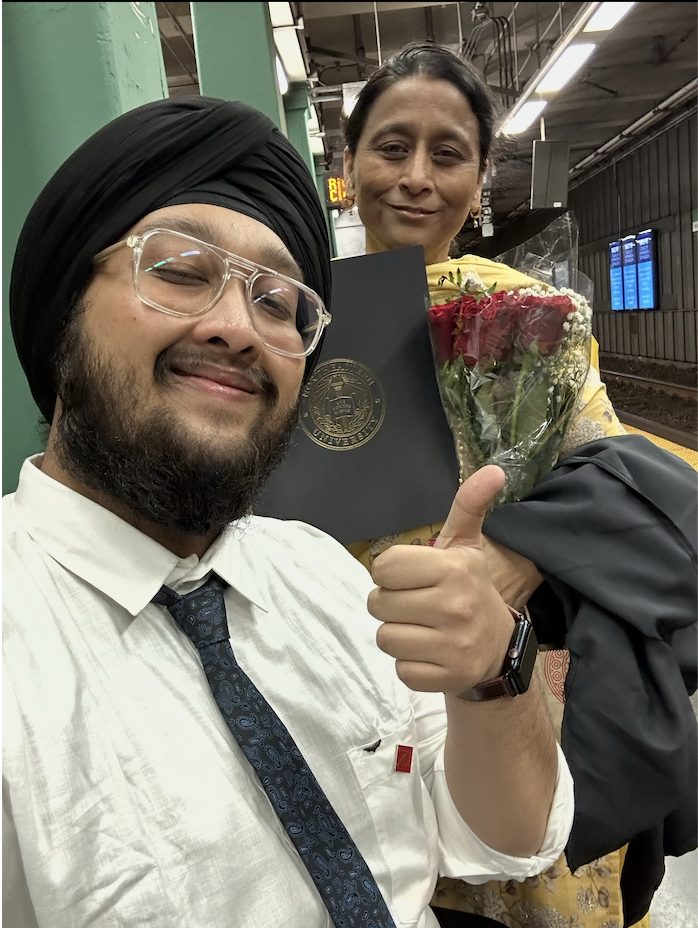 Dave in a suit and turban with his mom, holding a diploma folder and flowers, giving a thumbs up