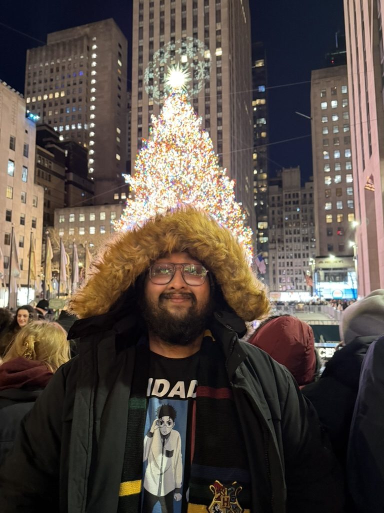 Dave in a fur-trimmed hood in front of a lit Christmas tree and tall buildings at night